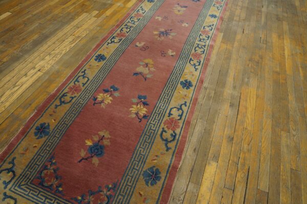 A long runner rug with a dusty rose field and blue and gold floral pattern sits on worn hardwood flooring.