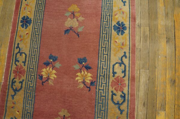 Reddish-pink runner rug featuring yellow and blue florals, detailed tan and blue borders, resting on rustic wood planks.