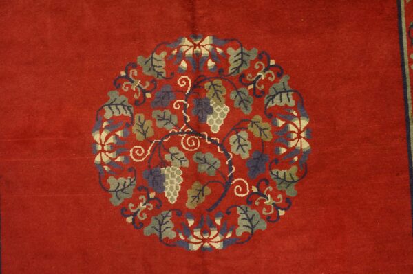 Red rug field with a central circular medallion of green and blue grapevines and fruit clusters.