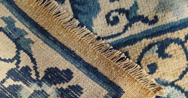 Two blue and tan patterned rug sections, one geometric and one scrolling, meet at a diagonal fringed edge.
