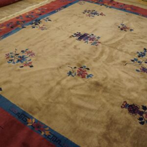 Shifting pile tan rug with scattered multicolored floral patterns and broad red and blue borders on wood.