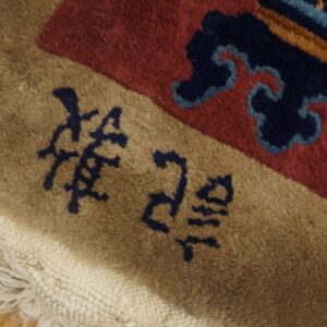 Tan rug border featuring dark blue script next to a red field with abstract blue patterns and white fringe.