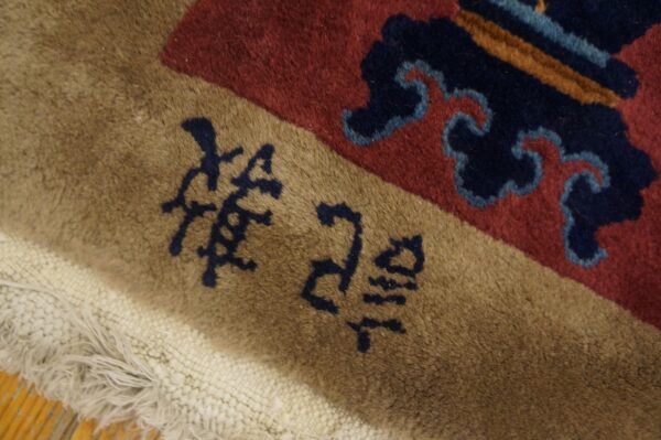 Tan rug border featuring dark blue script next to a red field with abstract blue patterns and white fringe.