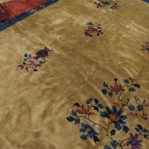 A lustrous tan rug features sparse blue, red, and purple floral motifs bordering a wide patterned red band.