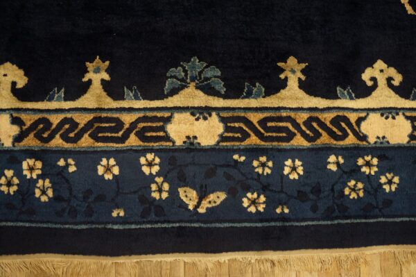 Close-up of a dark blue rug border featuring tan floral scrolls, geometric bands, and fringe on wood flooring.