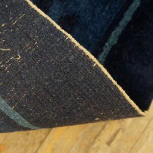 Rug corner curled up, showing a dark blue woven backing with a stripe and tan edge on wood planks.