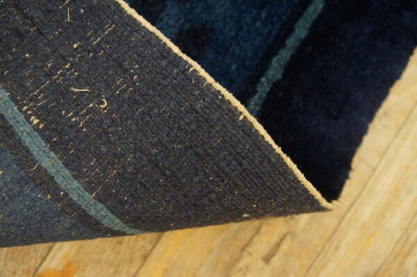 Rug corner curled up, showing a dark blue woven backing with a stripe and tan edge on wood planks.