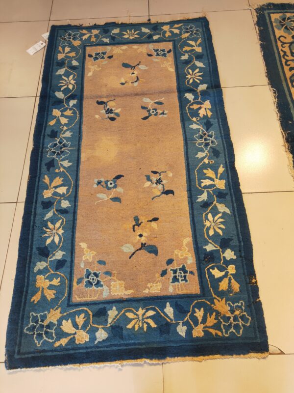 Tan field runner rug with blue borders featuring yellow and light blue floral vine designs on tiled flooring.