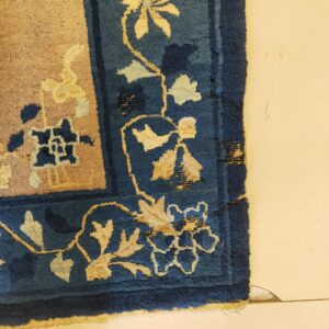 A close-up shows a deep blue rug border with cream floral motifs next to a tan field on light flooring.