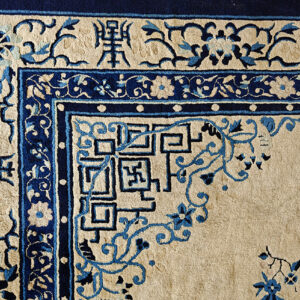 The corner of a high-pile beige rug with dark blue borders and traditional symbolic characters.