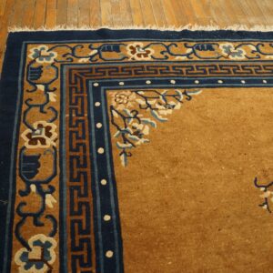 Golden brown rug corner with navy and tan scrolling borders and fringe visible on wood flooring.