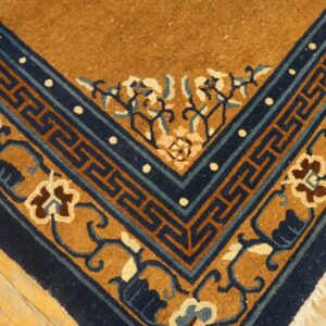 Tan rug corner featuring navy blue key fret and floral borders against light hardwood flooring.