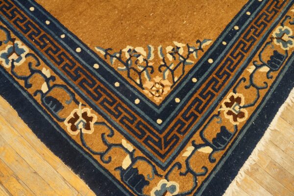 Tan rug corner featuring navy blue key fret and floral borders against light hardwood flooring.