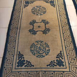 Low-pile tan rug with dark blue circular motifs and a Greek key border on white tile.