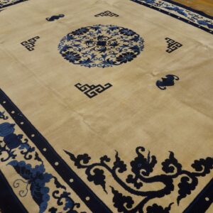 Tan area rug featuring navy blue floral borders, a central medallion, and stylized corner symbols on wood flooring.