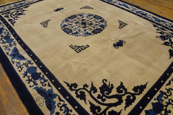 Tan area rug featuring navy blue floral borders, a central medallion, and stylized corner symbols on wood flooring.
