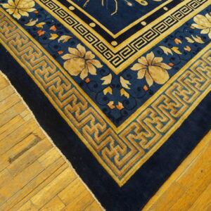 Dark blue rug with gold floral and fretwork borders rests on warm plank wood flooring.