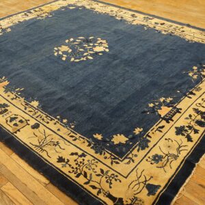 A square blue rug features a wide cream border with stylized floral and architectural patterns on wood floors.