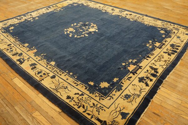 A square blue rug features a wide cream border with stylized floral and architectural patterns on wood floors.