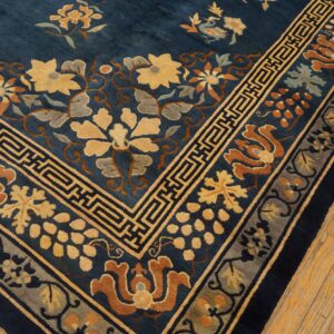Deep blue floral rug corner showing yellow, rust, and gray patterns with a fret border on light wood floors.
