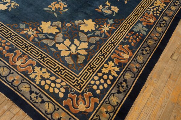 Deep blue floral rug corner showing yellow, rust, and gray patterns with a fret border on light wood floors.