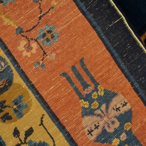 Woven detail of a rug with orange ground, featuring blue, gold, and pink geometric floral and butterfly motifs.