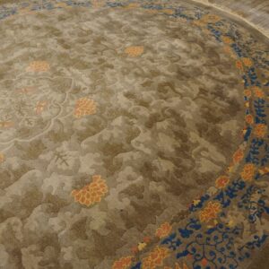Round, taupe rug featuring carved botanical patterns and a border with blue and orange floral accents on hardwood floors.