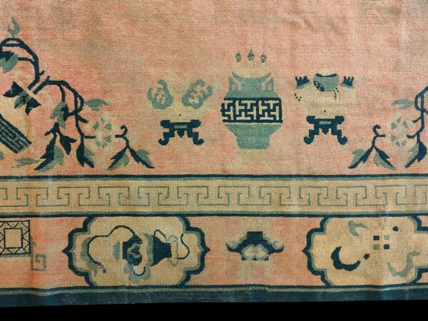 Low-pile rug detail showing a pink field with teal vase motifs, floral vines, and geometric borders.