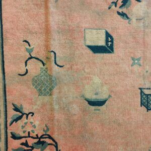 Faded pink rug detail showing teal and beige symbolic motifs, including vases and a Greek key border.