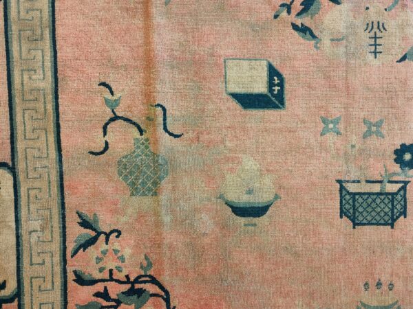 Faded pink rug detail showing teal and beige symbolic motifs, including vases and a greek key border.