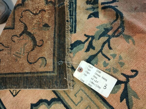 Muted peach rug with dark teal floral designs, showing the texture, visible fringe, and an attached sales tag.