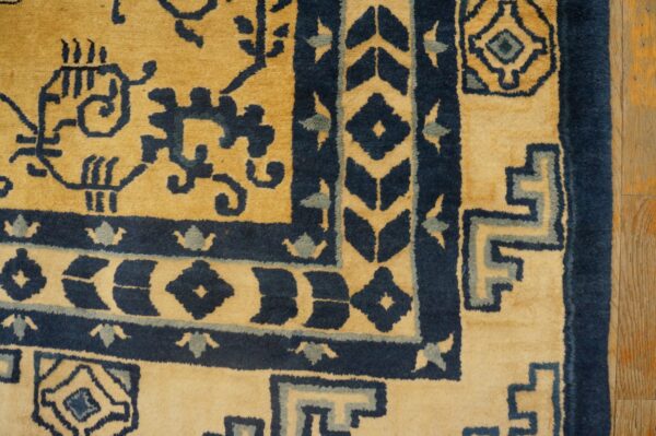 Corner of a gold and dark blue patterned rug showing geometric borders next to distressed wood flooring.