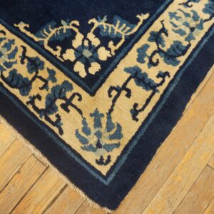 Corner of a navy blue and tan rug featuring a stylized floral border on worn, golden-toned wood flooring.
