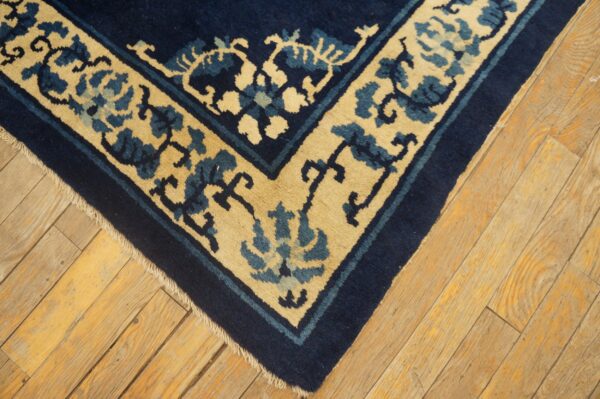 Corner of a navy blue and tan rug featuring a stylized floral border on worn, golden-toned wood flooring.
