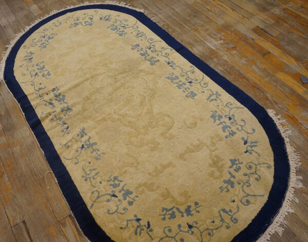 A tan oval rug with pale blue scrollwork and a navy border lies on distressed wooden flooring.