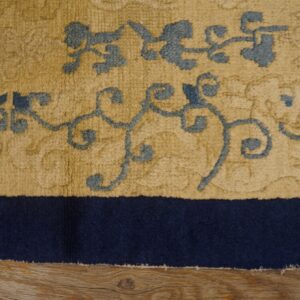 High-pile tan rug with slate blue scrolling patterns and a navy border shown partially on wood flooring.
