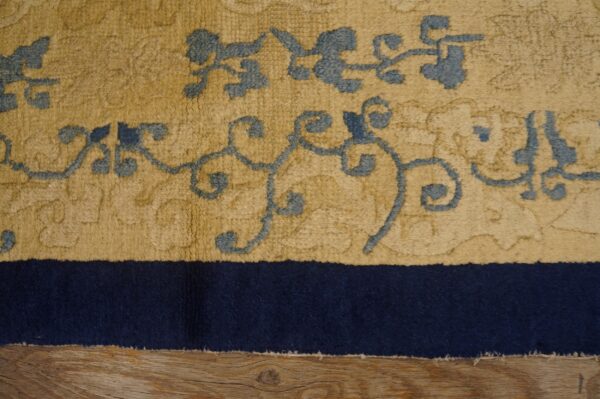 High-pile tan rug with slate blue scrolling patterns and a navy border shown partially on wood flooring.
