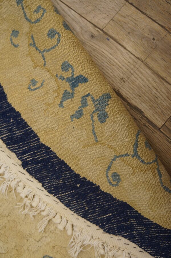 Textured tan and blue patterned rug with fringe, partially curled on light brown wooden flooring.