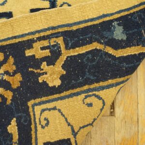 A curled rug corner features navy blue and golden geometric patterns on rustic light wood floors.