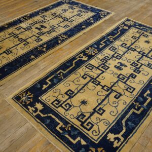 Two rectangular rugs with beige fields and navy blue geometric patterns lie on a rustic wood floor.