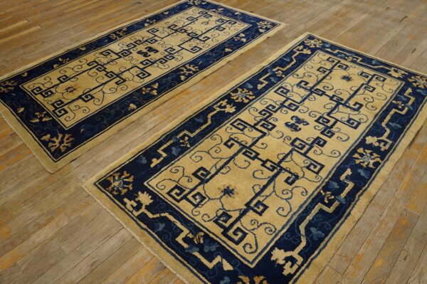 Two rectangular rugs with beige fields and navy blue geometric patterns lie on a rustic wood floor.