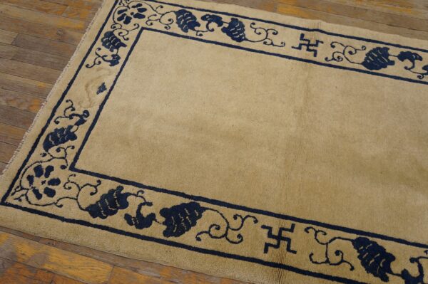 Tan rug with dark blue vine and swastika borders rests on distressed wooden plank flooring.