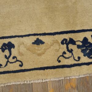 Tan rug with dark blue floral borders and visible fringe rests on rustic wood flooring.