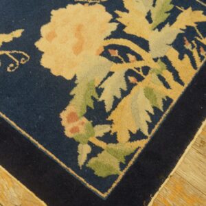 Dark blue square rug with large pale yellow and green floral motifs rests on distressed light wooden flooring.
