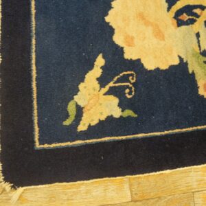 Thick pile, navy blue square rug displaying large yellow and green floral designs sits on light golden wood planks.