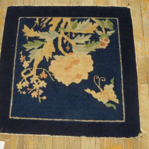 Navy blue square rug featuring a large floral design in pale yellow and green rests on rustic wood planks.