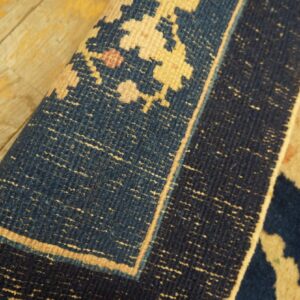 Deep blue and light tan patterned square rug detail, showing the border curled on wood floorboards.