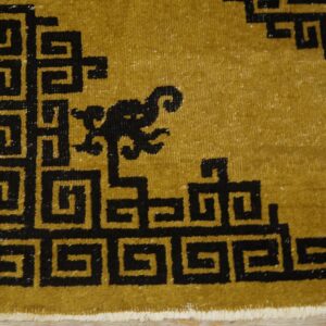 Mustard-colored rug corner with a black meander pattern and a small stylized motif on wood flooring.