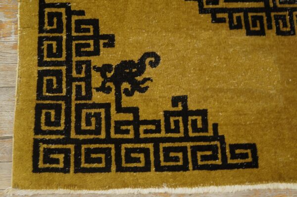 Mustard-colored rug corner with a black meander pattern and a small stylized motif on wood flooring.
