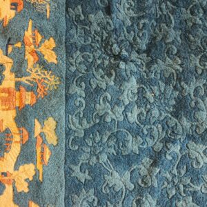 Close view of a blue rug with a carved pile floral field and an orange patterned border.